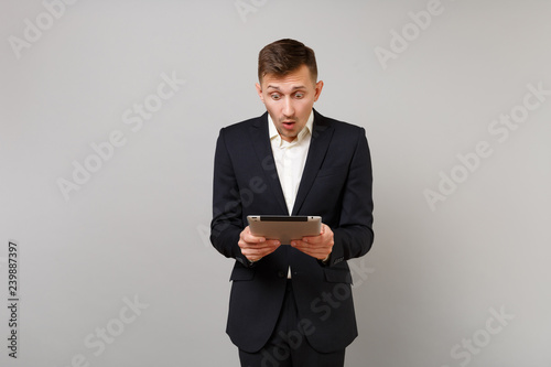 Shocked young business man in suit keeping mouth open, looking surprised holding, using tablet pc computer isolated on grey background. Achievement career wealth business concept. Mock up copy space.