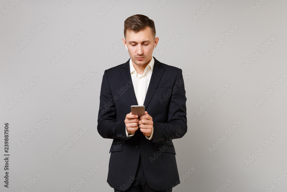 Young business man in suit with wireless earphones listening music, using mobile phone, typing sms message isolated on grey background. Achievement career wealth business concept. Mock up copy space. Young business man in suit with wireless earphones listening music, using mobile phone, typing sms message isolated on grey background. Achievement career wealth business concept. Mock up copy space.
