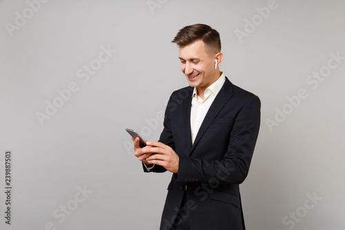 Smiling young business man in classic black suit with wireless earphones listening music using mobile phone isolated on grey background. Achievement career wealth business concept. Mock up copy space.