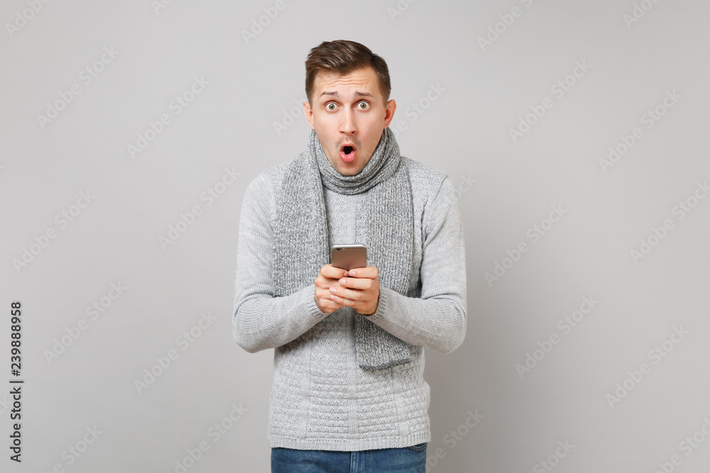 Shocked young man in gray sweater, scarf using mobile phone typing sms message, keeping mouth open isolated on grey background. Healthy fashion lifestyle, people sincere emotions, cold season concept. Shocked young man in gray sweater, scarf using mobile phone typing sms message, keeping mouth open isolated on grey background. Healthy fashion lifestyle, people sincere emotions, cold season concept.