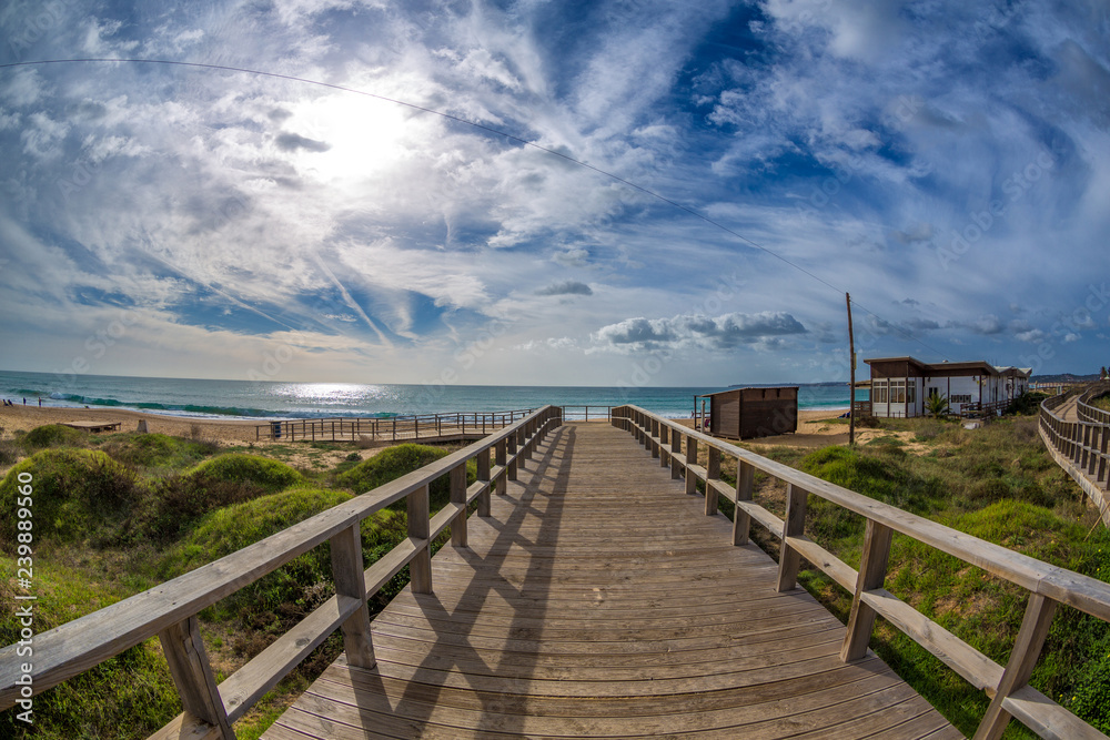 Fototapeta premium Wooden walkways lead to water in famous beach of Tres Irmaos in Alvor, Portimão, Algarve, Portugal, Europe. Praia dos Tres Irmaos. The sun shines through the clouds, the waves roll on the sand.