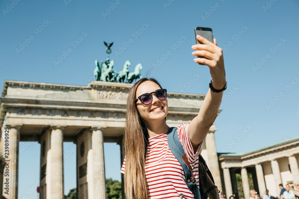 Young beautiful positive girl makes selfie against the background of the Brandenburg Gate in Berlin in Germany or takes pictures of sights.