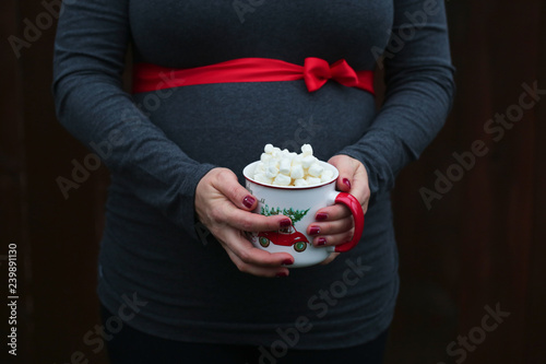 Pregnancy, Christmas and People Concept - close up of Pregnant Woman with Red ribbon over, holding Cup with Hot Coco and  Mushmellow  photo