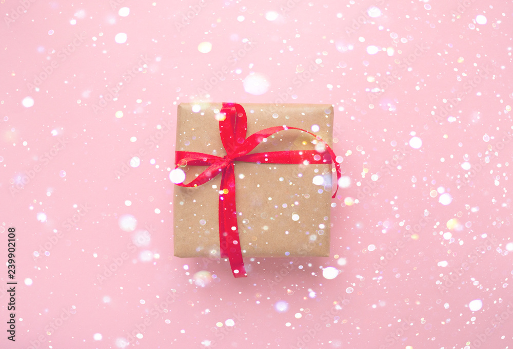 Gift box wrapped in brown colored craft paper and tied with red bow on pink background with garland lights bokeh effect. Gift box wrapped in brown colored craft paper and tied with red bow on pink background with garland lights bokeh effect.