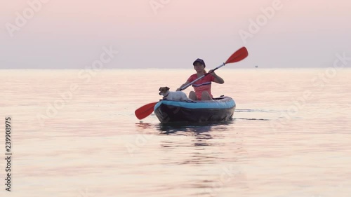 Varna, Bulgaria, mid December 2018. Young woman wearing a cap paddles on an inflatable kayak over the calm surface of the sea against the sky in the light of a pink dawn. Dog Jack Russell with kayak photo