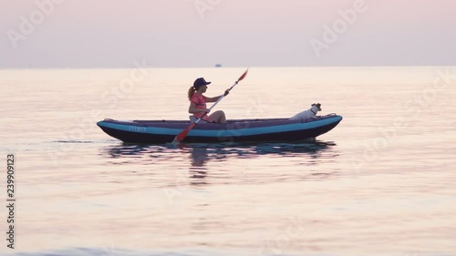 Varna, Bulgaria, mid December 2018. Young woman wearing a cap paddles on an inflatable kayak over the calm surface of the sea against the sky in the light of a pink dawn. Dog Jack Russell with kayak photo