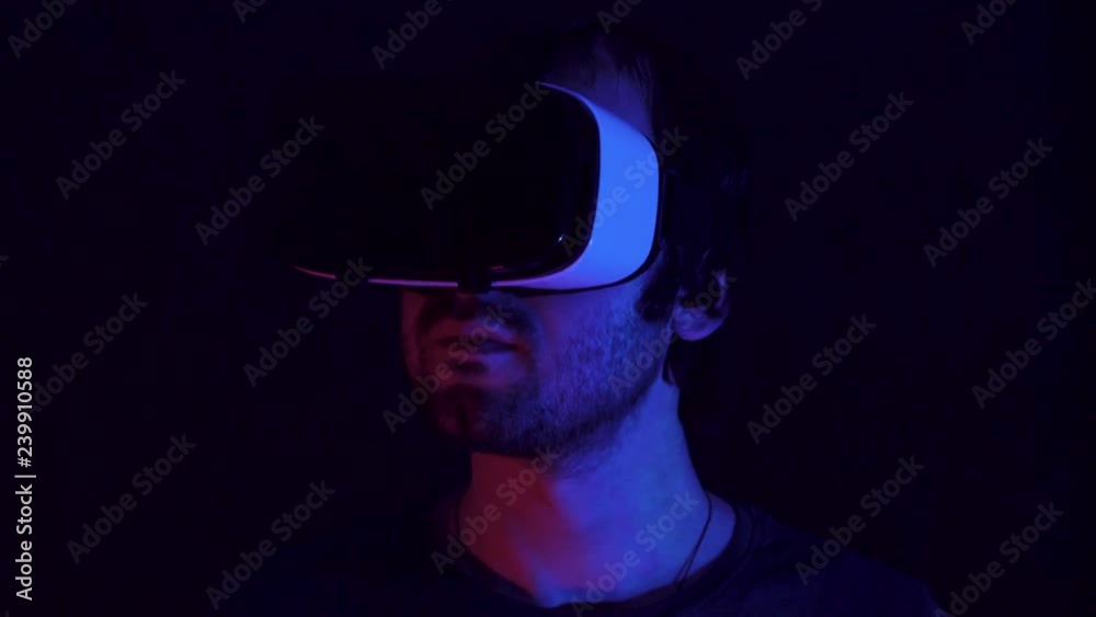 Young man in VR headset looks around and wonders how amazing. Virtual reality helmet on black technology background. Modern fluorescent light Young man in VR headset looks around and wonders how amazing. Virtual reality helmet on black technology background. Modern fluorescent light