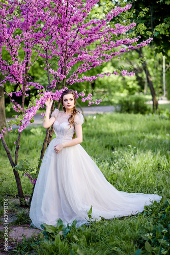 Luxurious and stylish bride poses in the garden among purple flowers. Happy bride with beautiful and long hair. An important day in life, strong emotions.