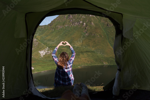 Personal perspective of a male camper in tent in the swiss alps with a young woman doing hand heart shape in front of him © NDStock