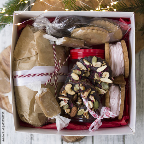 White box with Christmas sweets - Stollen, Cookies, a jar of salted caramel, toffee, marshmallows, chocolate rings with nuts, homemade Christmas gift, Christmas treeon white wooden table. From above photo