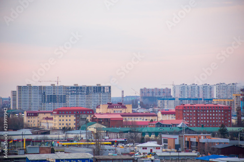 Landscape of evening city with high-rise buildings under construction, factories and pipes with smoke at sunset under boundless cloudy sky