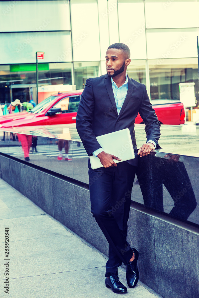 Obraz premium Young African American Businessman with beard, afro short hair, traveling in New York, wearing black suit, leather shoe, holding laptop computer, standing on street in Middletown of Manhattan, looking