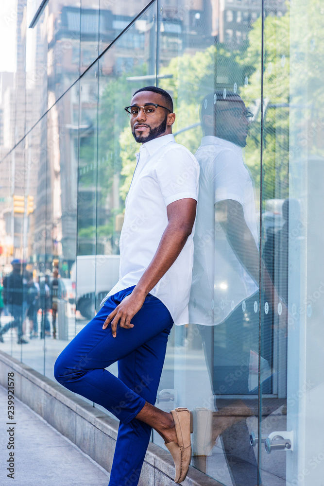 Young African American Man with beard, wearing white short sleeve shirt, blue pants, casual shoes, glasses, standing against glass wall with reflections on street in Middletown of Manhattan, New York Young African American Man with beard, wearing white short sleeve shirt, blue pants, casual shoes, glasses, standing against glass wall with reflections on street in Middletown of Manhattan, New York