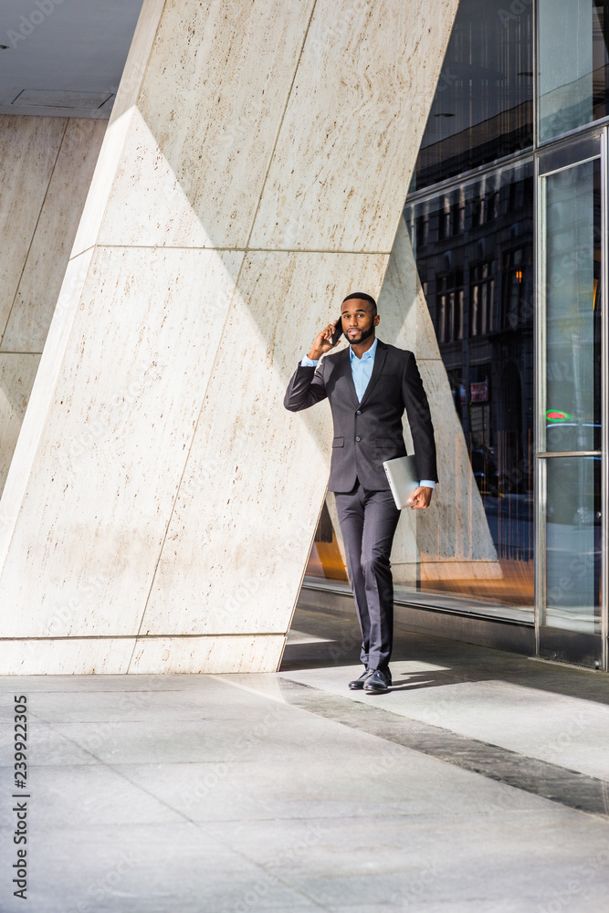 African American businessman with beard talking on cell phone, traveling, working in New York, wearing black suit, leather shoes, carrying laptop computer, walking out from office building to street