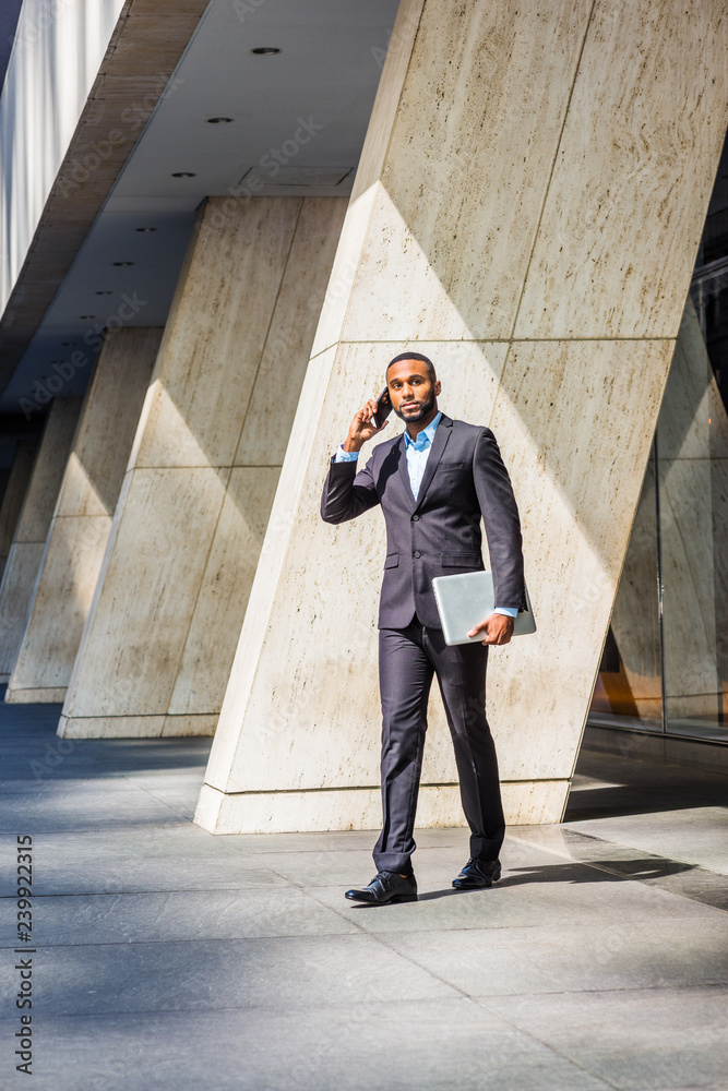 African American businessman with beard talking on cell phone, traveling, working in New York, wearing black suit, leather shoes, carrying laptop computer, walking out from office building to street