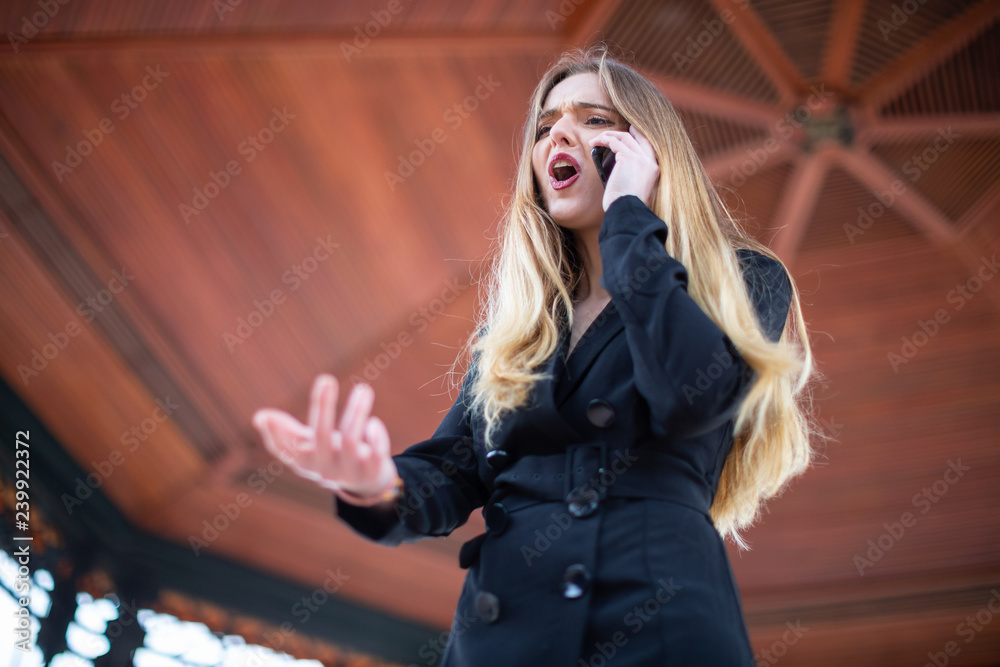 Beautiful young female in elegant outfit screaming and gesturing while being fired from work by phone in park