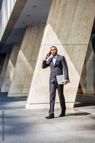 Obraz na płótnie African American businessman with beard talking on cell phone, traveling, working in New York, wearing black suit, leather shoes, carrying laptop computer, walking out from office building to street w salonie
