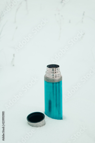 Close-up mint silver thermos with coffee or tea in snow in snowy forest. Winter holidays, hot drinks concept.