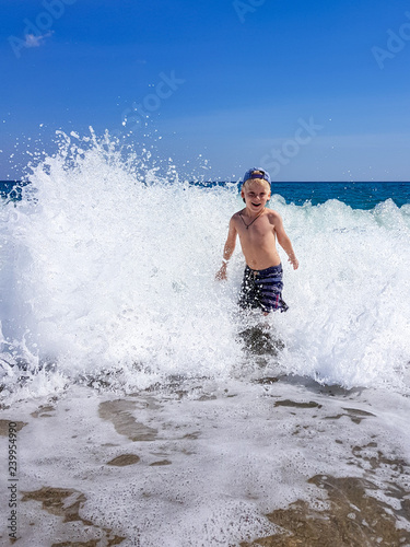 A little blond boy is playing on the seashore, laughing, running away from the waves against the background of huge waves and splashes, Calabria, Italy