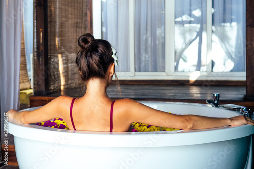 Back of woman in a bathtub with tropical exotic flowers in the water and flower white plumeria in her hair.girl in eco hotel taking bath in the spa salon summer vacation in a deluxe room ,skin care