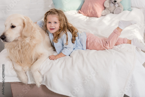 high angle view of adorable kid hugging golden retriever on bed in children room and looking at camera