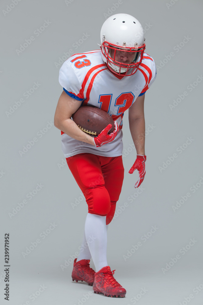 Full length shot of american football sportsman player wearing helmet posing with rugby ball isolated on grey background
