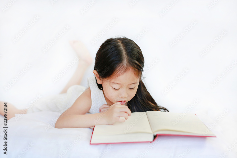 Beautiful little Asian child girl reading hardcover book lying with pillow on white background. Beautiful little Asian child girl reading hardcover book lying with pillow on white background.