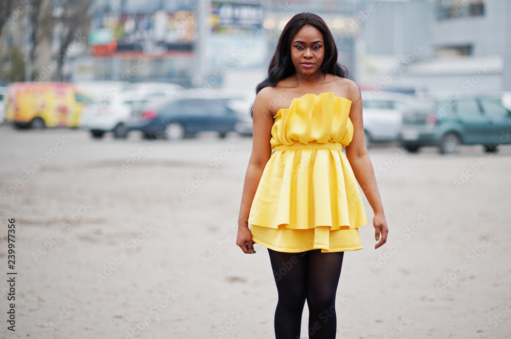 Stylish african american woman at yellow dress posed outdoor car parking.