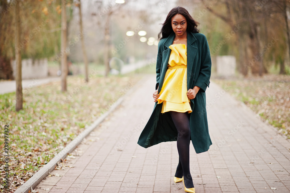 Fototapeta premium Stylish african american woman at green coat and yellow dress posed against autumn park.