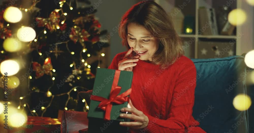 Portrait shot of the attractive and happy caucasian woman sitting at the x-mas tree in the living room and opening a present box from inside of which there is a strong shining light. Portrait shot of the attractive and happy caucasian woman sitting at the x-mas tree in the living room and opening a present box from inside of which there is a strong shining light.