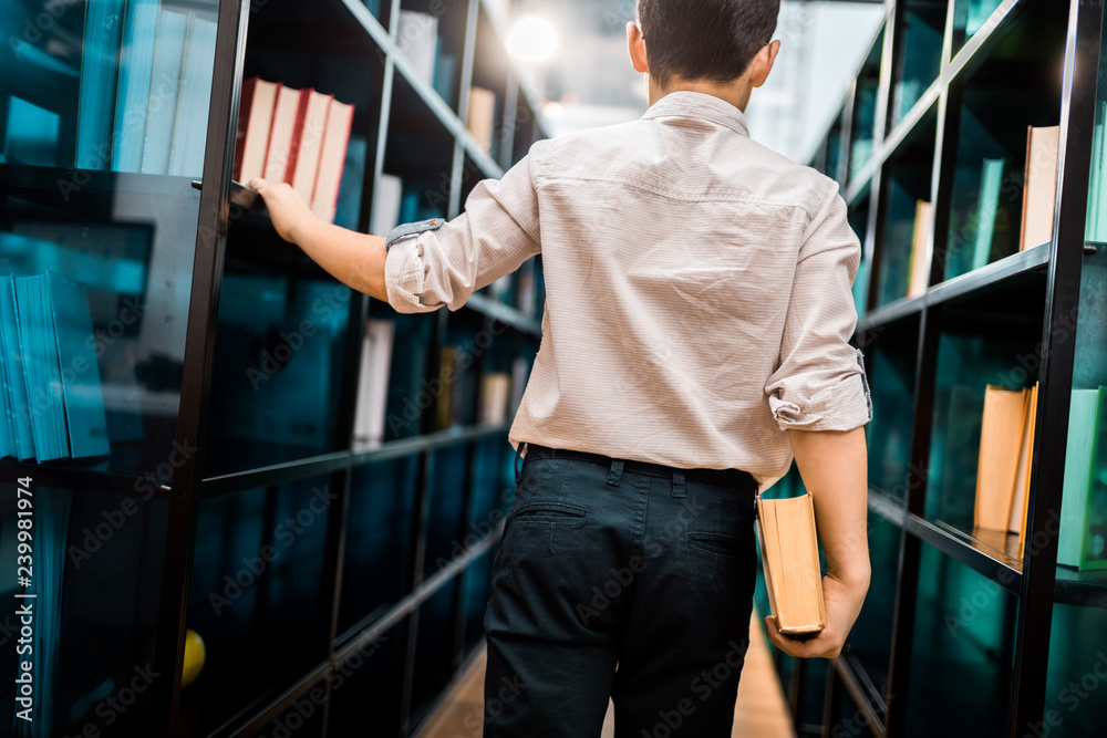 back view of boy holding book and walking between bookshelves in library