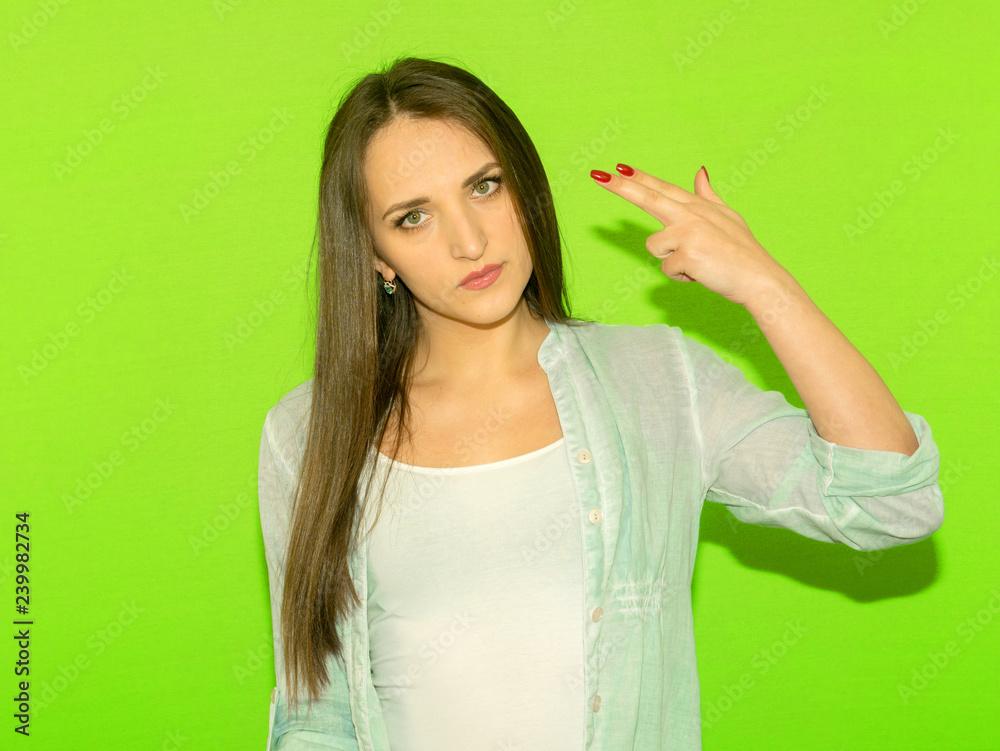 Studio portrait of a tired girl shooting herself doing a gun mark with her finger .Isolated on a green background. Human facial expressions Studio portrait of a tired girl shooting herself doing a gun mark with her finger .Isolated on a green background. Human facial expressions