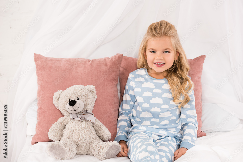 cute smiling child sitting on bed with teddy bear and pillows