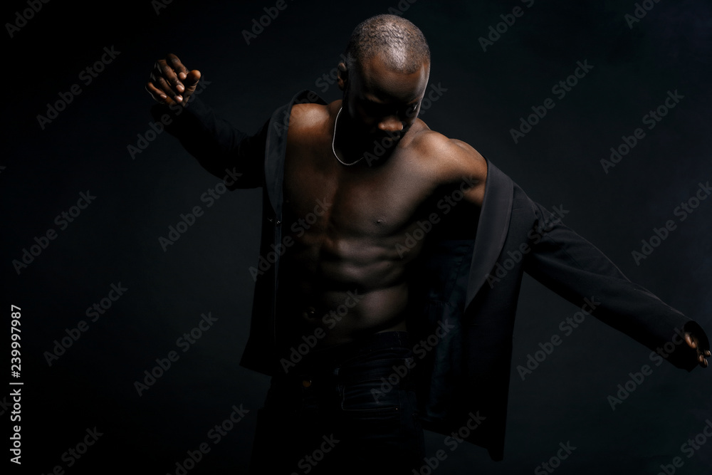 Dark key portrait of sexy dancer man topless, with jacket on shoulders and chainlet on neck. Studio shot, black background