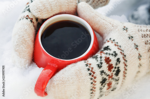 Hands in woolen mittens hold a red mug with hot tea or coffee, standing on the snow. Picnic on a frosty Sunny day. The concept of warming on a winter walk