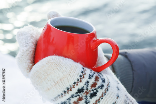 Hands in woolen mittens hold a red mug with hot tea or coffee. Picnic on a frosty Sunny day on the snowy Bank of the river or sea. The concept of warming on a winter walk photo