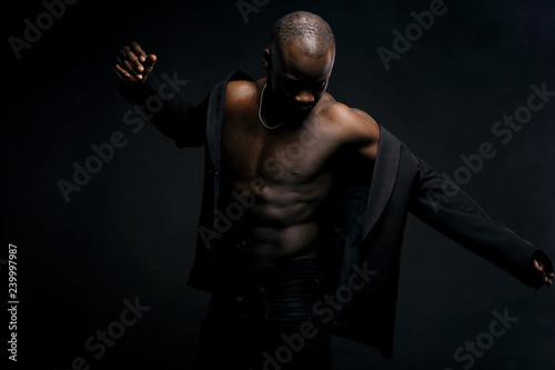 Dark key portrait of sexy dancer man topless, with jacket on shoulders and chainlet on neck. Studio shot, black background