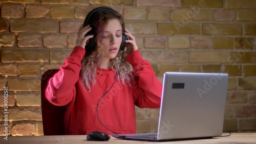 Portrait of young girl in red hoodie putting on her headphones and watching into laptop on bricken wall background. photo