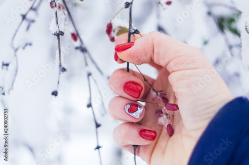 Valentine day nail art design manicure. Female hand with bright red heart manicure holding red berries on a winter background. Trendy red nails with design. Red Nail polish. Selective focus