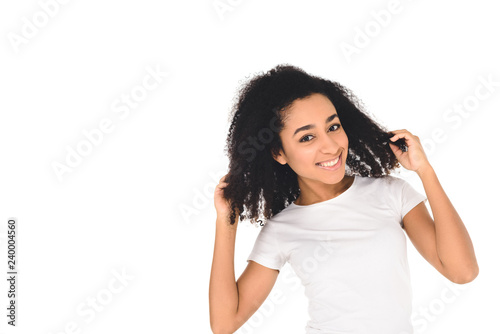 attractive african american girl touching hair and smiling at camera isolated on white