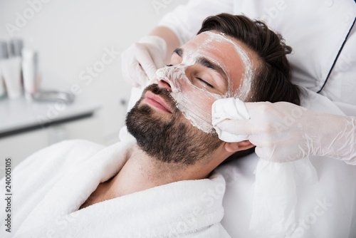 Obraz na plátně Relaxed man closing eyes and cosmetologist wiping out mask