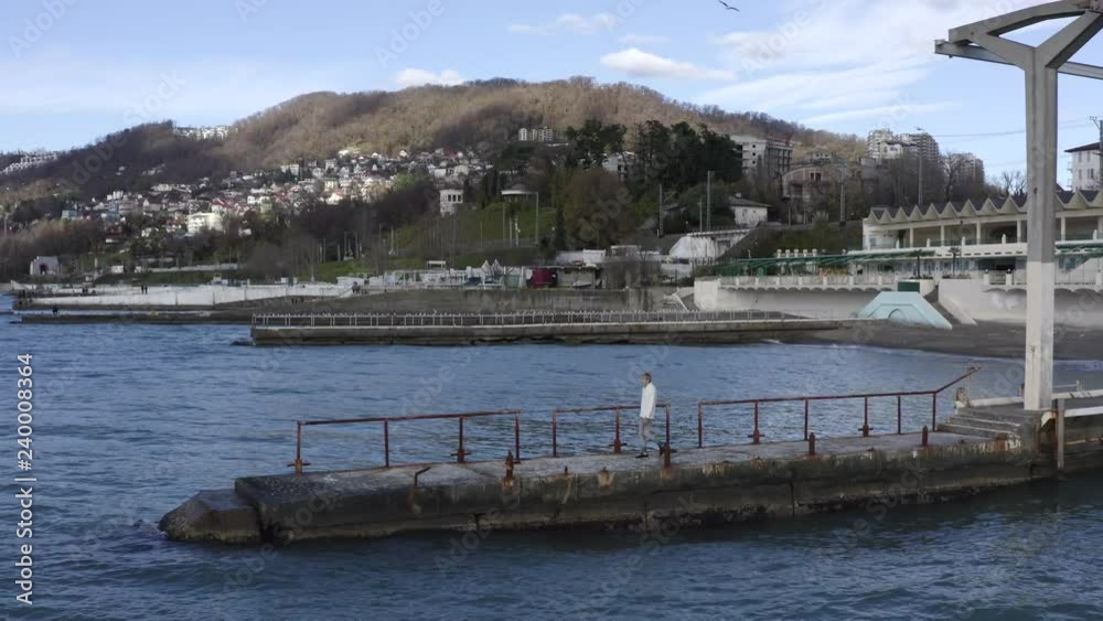 Man walking on pier outstretching arms and enjoying beautiful landscape drone view. Young man standing on sea pier and looking up. Embankment of marine city, building and highlands background Man walking on pier outstretching arms and enjoying beautiful landscape drone view. Young man standing on sea pier and looking up. Embankment of marine city, building and highlands background