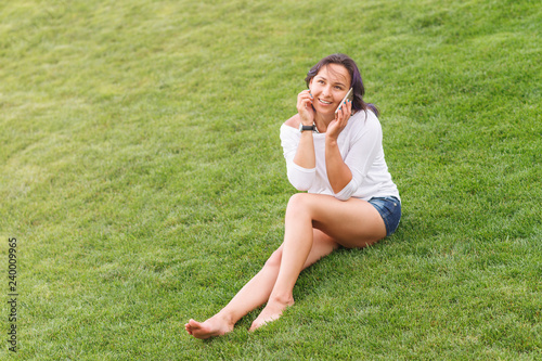 Happy beautiful young woman sitting on grass and using smartphone © mikhail_b_azarov