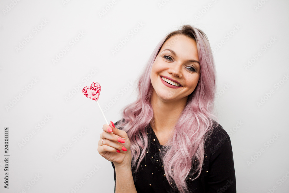 Lovely charming girl with a Lollipop in the form of heart. Portrait of a young woman with pink hair and pink candy Lovely charming girl with a Lollipop in the form of heart. Portrait of a young woman with pink hair and pink candy