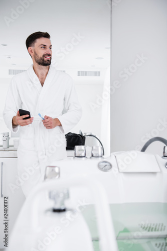 Waist up of smiling man in white bathrobe standing in the bathroom