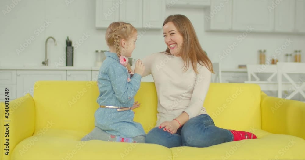 Cheerful elementary age daughter whispering gossips to surprised beautiful mother while sitting on sofa in domestic room. Smiling little girl revealing secrets to charming mom while relaxing at home. Cheerful elementary age daughter whispering gossips to surprised beautiful mother while sitting on sofa in domestic room. Smiling little girl revealing secrets to charming mom while relaxing at home.