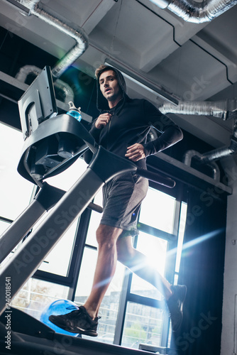 low angle view of handsome sportsman in hoodie exercising on treadmill in gym and looking at camera