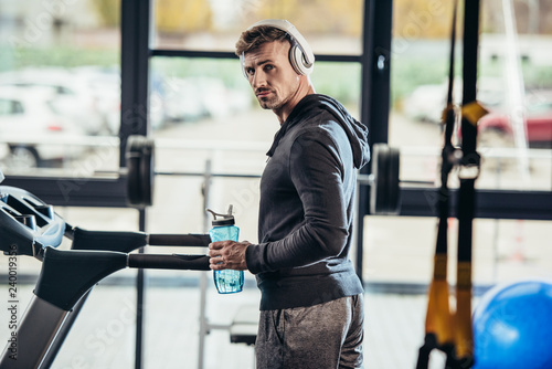 portrait of handsome sportsman in hoodie standing near treadmill with sport bottle in gym and looking at camera