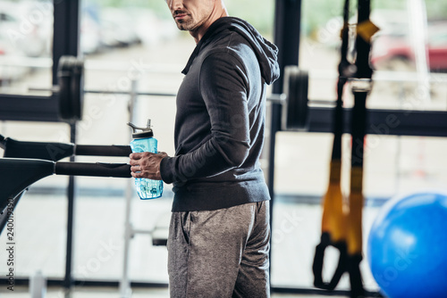 cropped image of sportsman in hoodie standing near treadmill with sport bottle in gym