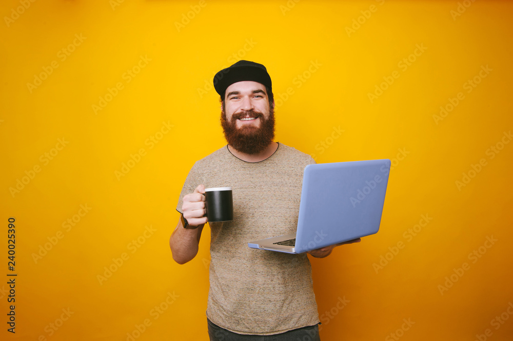 Cheerful bearded man looking at the camera holding a cup of coffee or tea and holding laptop Cheerful bearded man looking at the camera holding a cup of coffee or tea and holding laptop